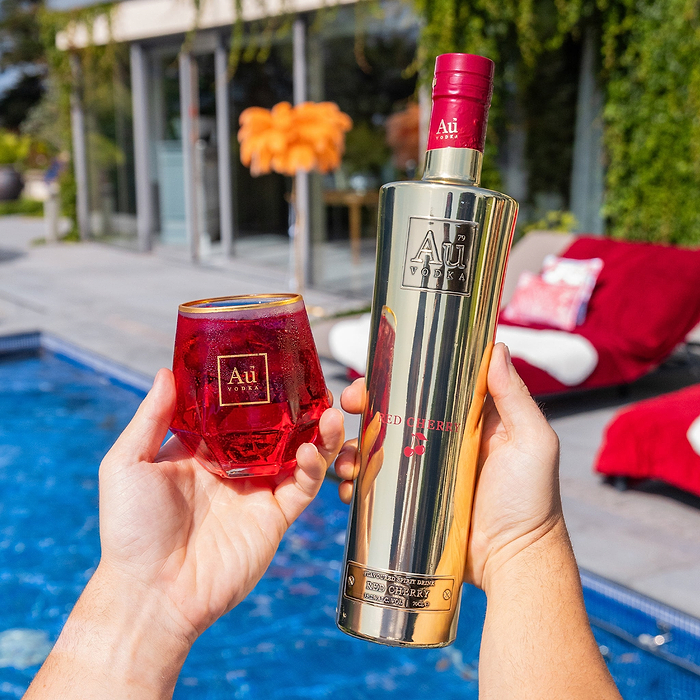 Hands holding a bottle of Au Vodka Red Cherry and a matching glass of red cocktail by a swimming pool