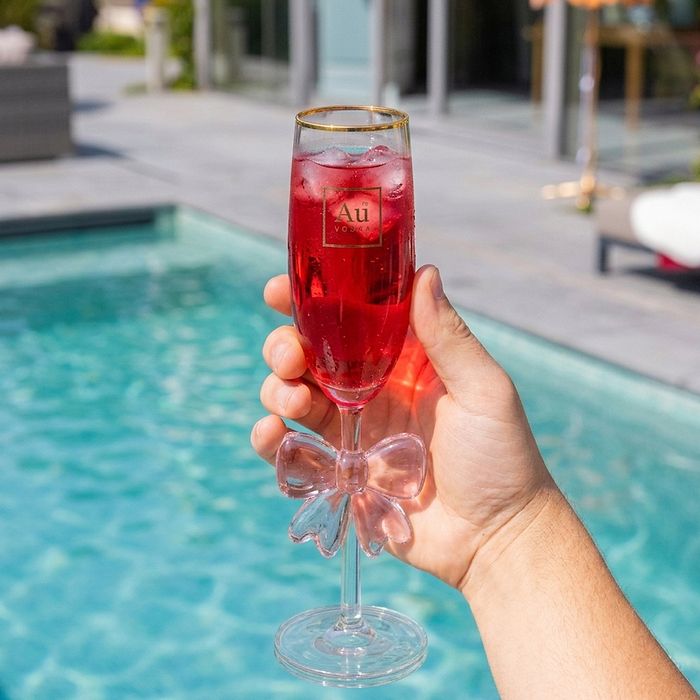 Hand holding an Au Vodka USA glassware flute with a bow detail, filled with a red drink and ice by a pool