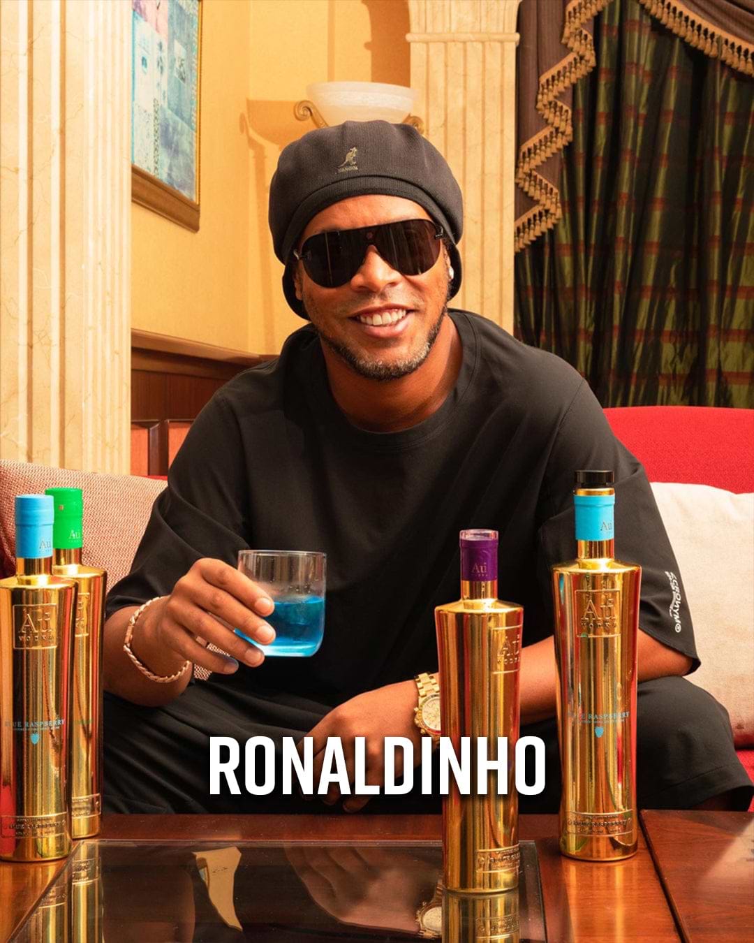 Ronaldinho wearing sunglasses and a black beanie, holding a glass of blue liquid, with Au Vodka bottles