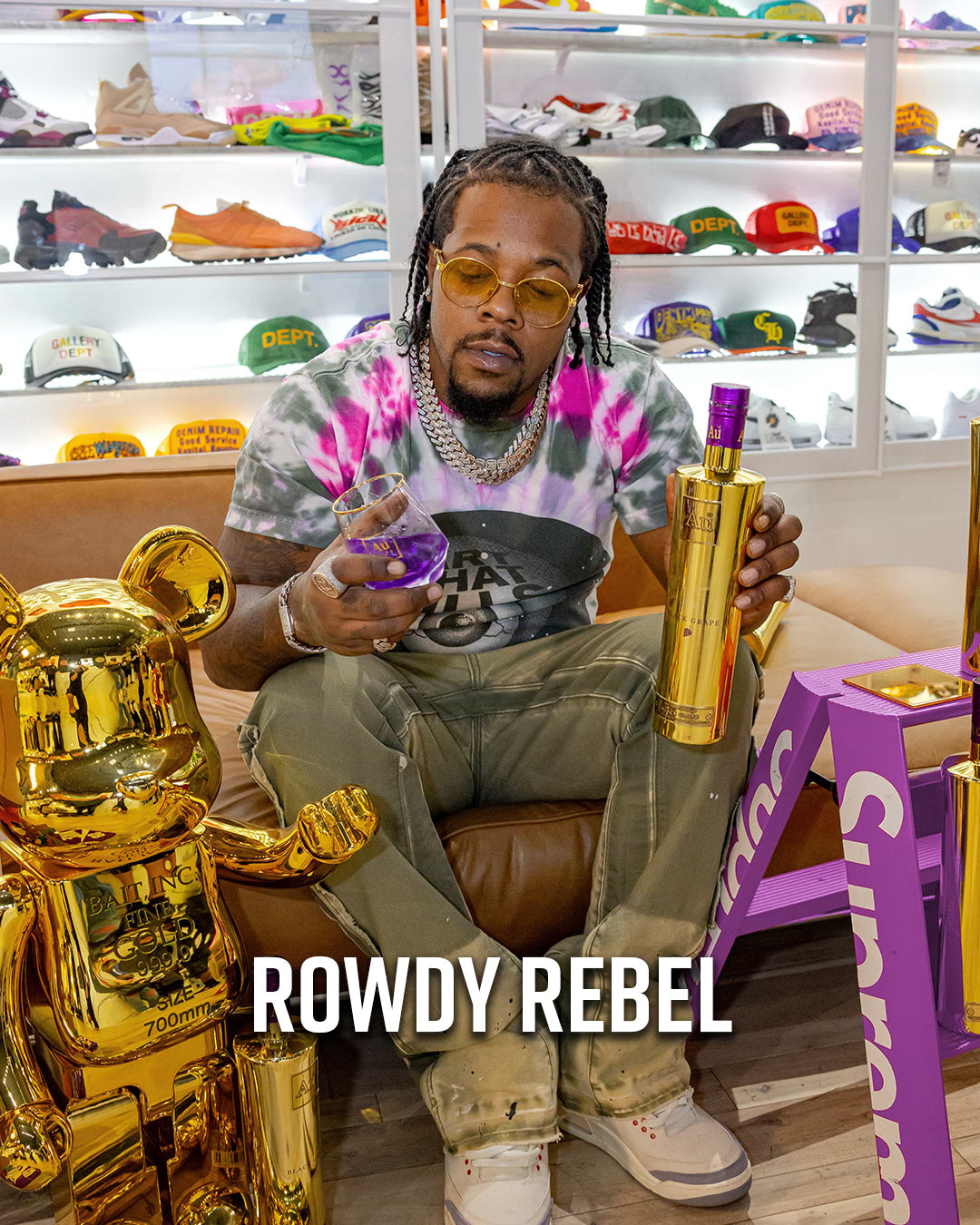 Rowdy Rebel holding a bottle of purple liquid and a glass, with a gold bearbrick figure beside him