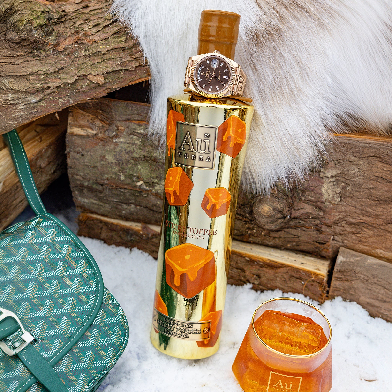 Au Vodka Sticky Toffee bottle with a luxury watch on top next to a Goyard bag and a glass of vodka on ice