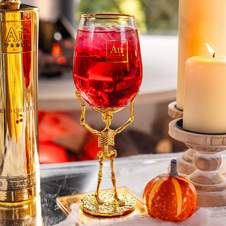 Au Vodka Red Cherry bottle next to a gold skeleton stemware glass filled with red drink and a small pumpkin
