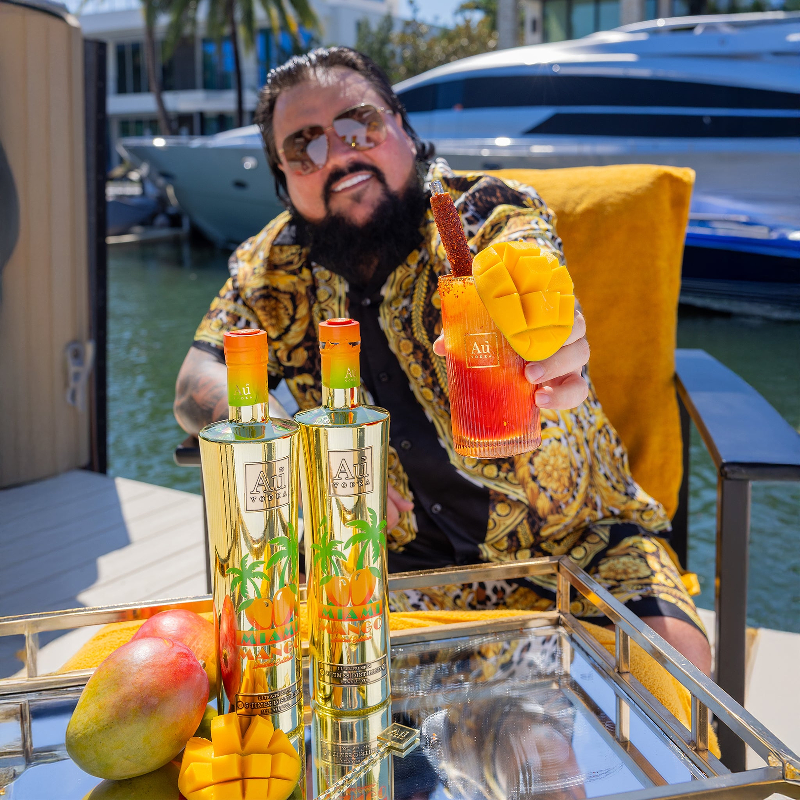 Man in sunglasses holding a mango cocktail, with two Au Vodka Miami Mango bottles and fresh mangoes