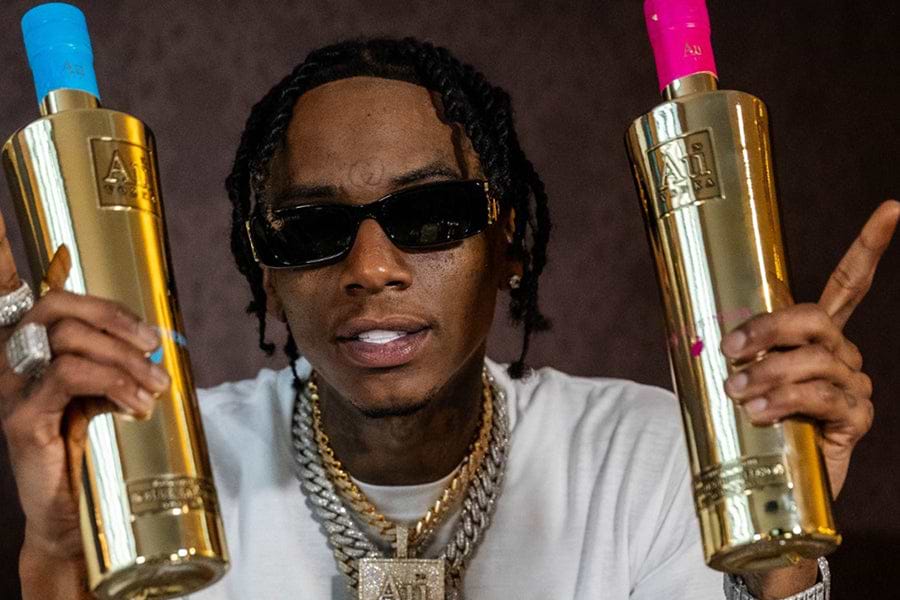 Man in sunglasses holding two gold Au Vodka bottles, one with a blue cap and one with a pink cap