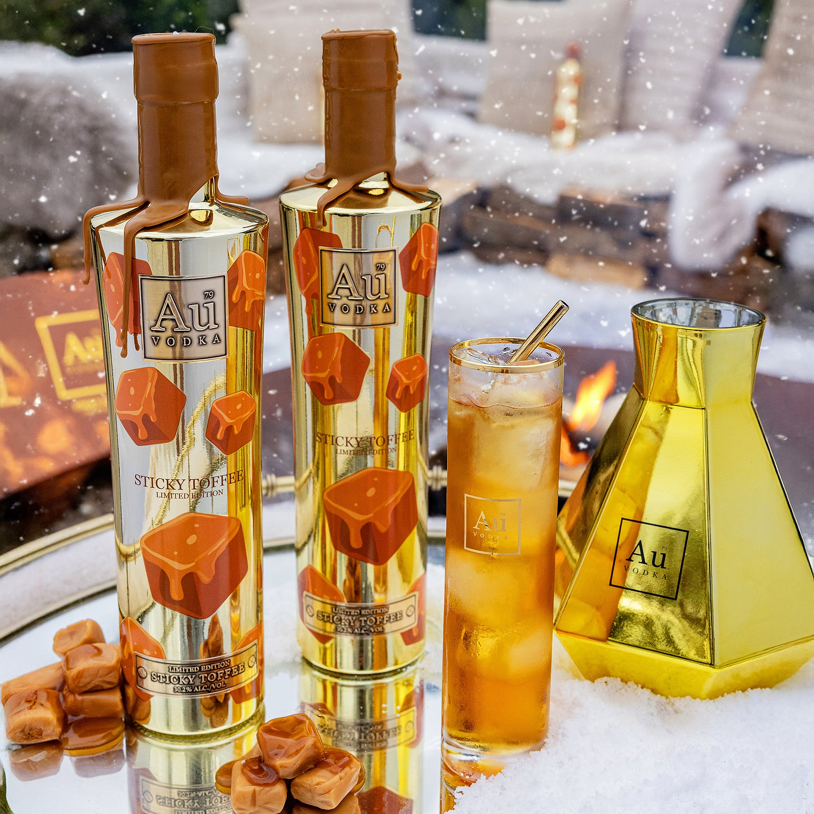 Two bottles of Au Pairs Sticky Toffee pudding with a glass of iced drink and caramels on a snowy background.