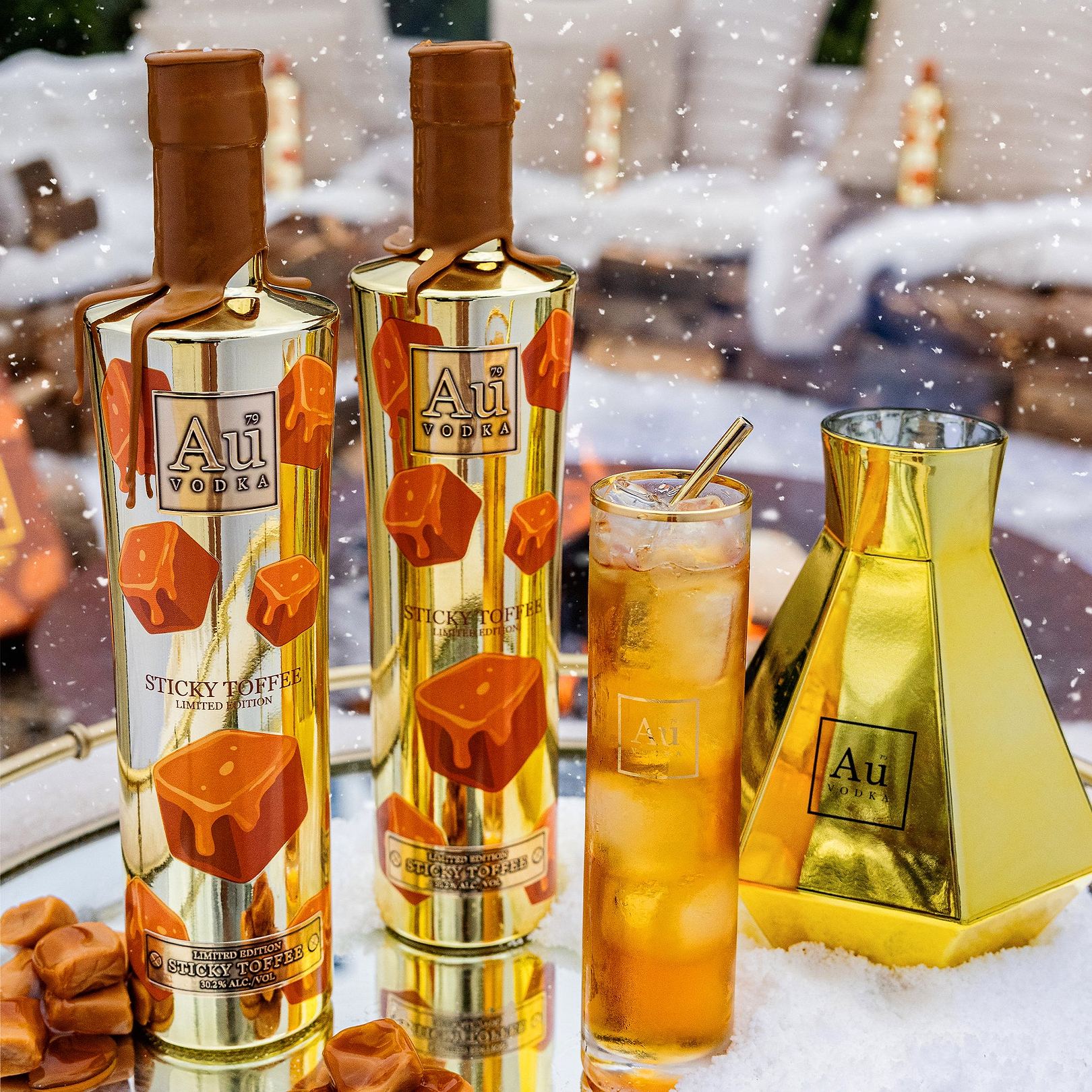 Two Au Vodka Sticky Toffee bottles with a cocktail and gold shaker in a snowy setting