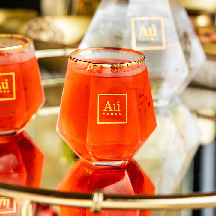 Red cocktail in a faceted Au Vodka glass with gold rim on a reflective surface