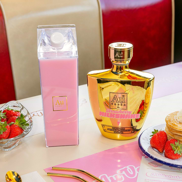 Clear carton of pink cocktail next to a gold Au Vodka bottle and strawberries