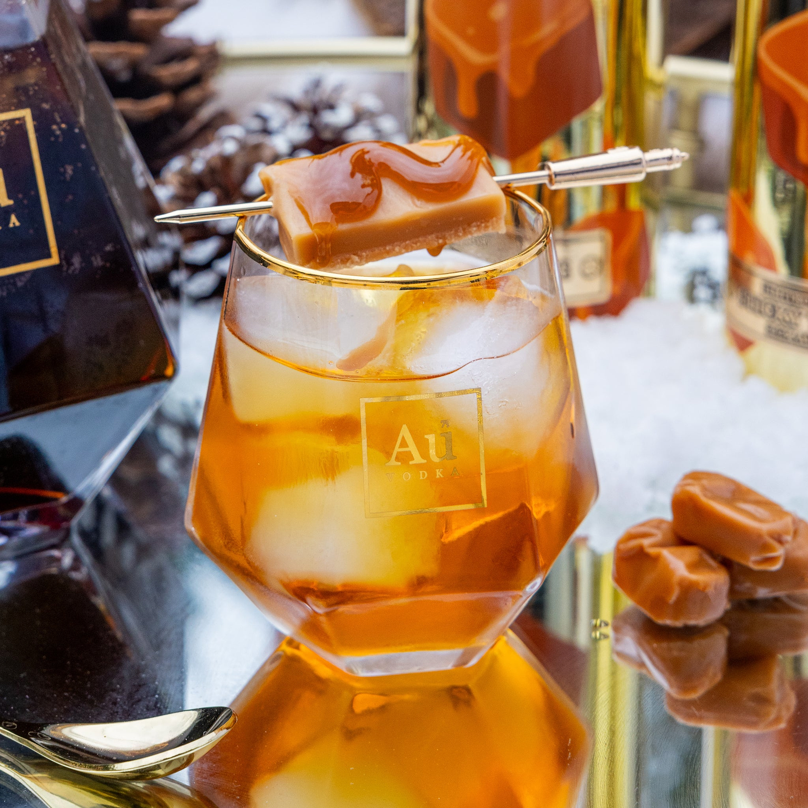 Glass of Au Vodka Sticky Toffee with a caramel square on a skewer, surrounded by bottles and caramel candies.