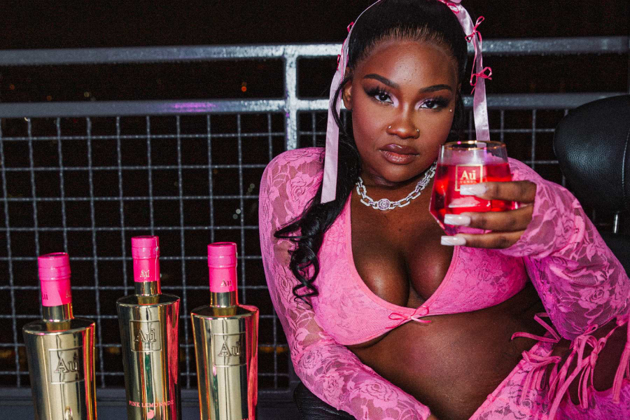 Woman in pink lace top holding a glass of Au Vodka Pink Lemonade, with three bottles on the left