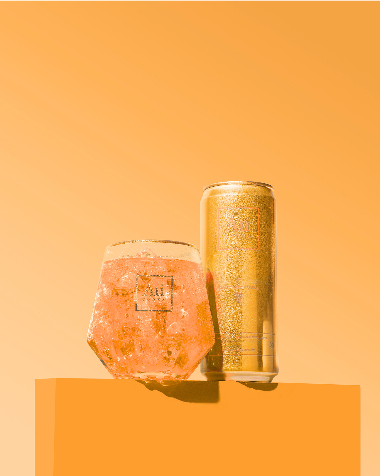 Au Vodka gold can and glass of orange drink with ice on orange pedestal