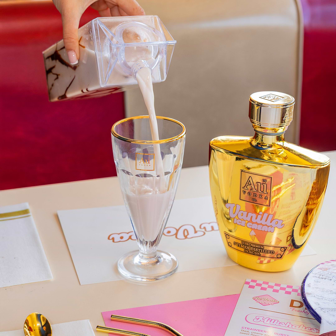 Hand pouring chocolate drink from a clear carton into a glass next to Au Vodka bottle