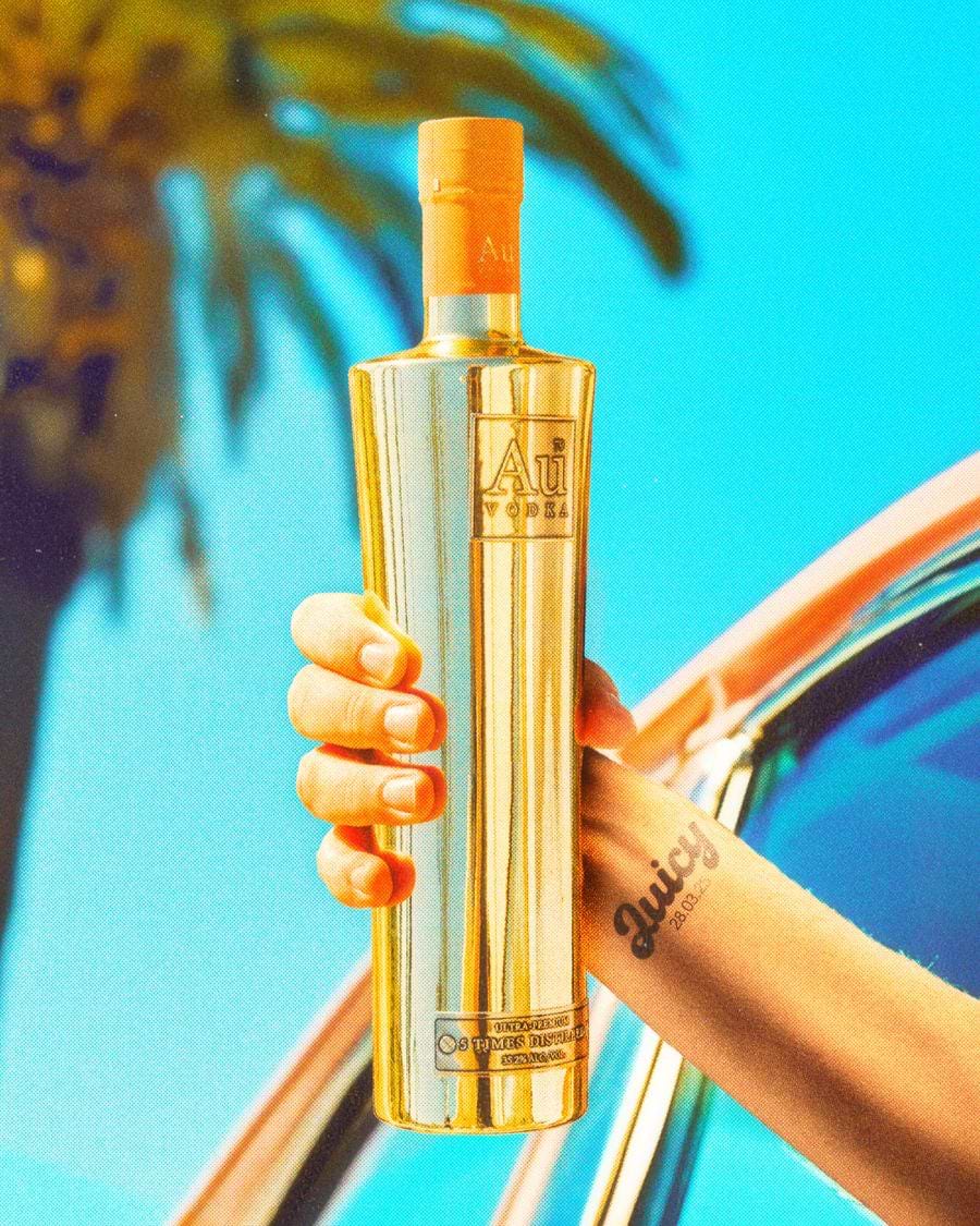 Hand holding a gold Au Vodka bottle against a bright blue sky and palm tree background