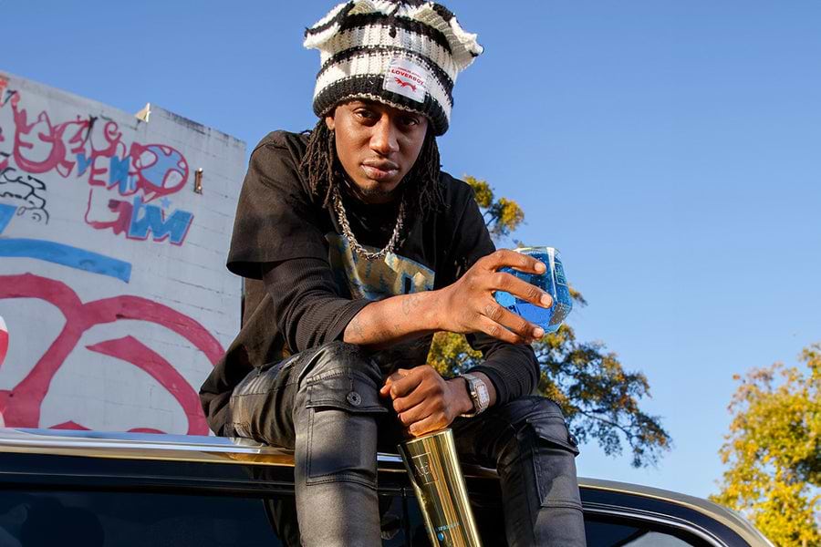 Man in a black and white striped beanie and black clothing holding a blue drink and a gold bottle