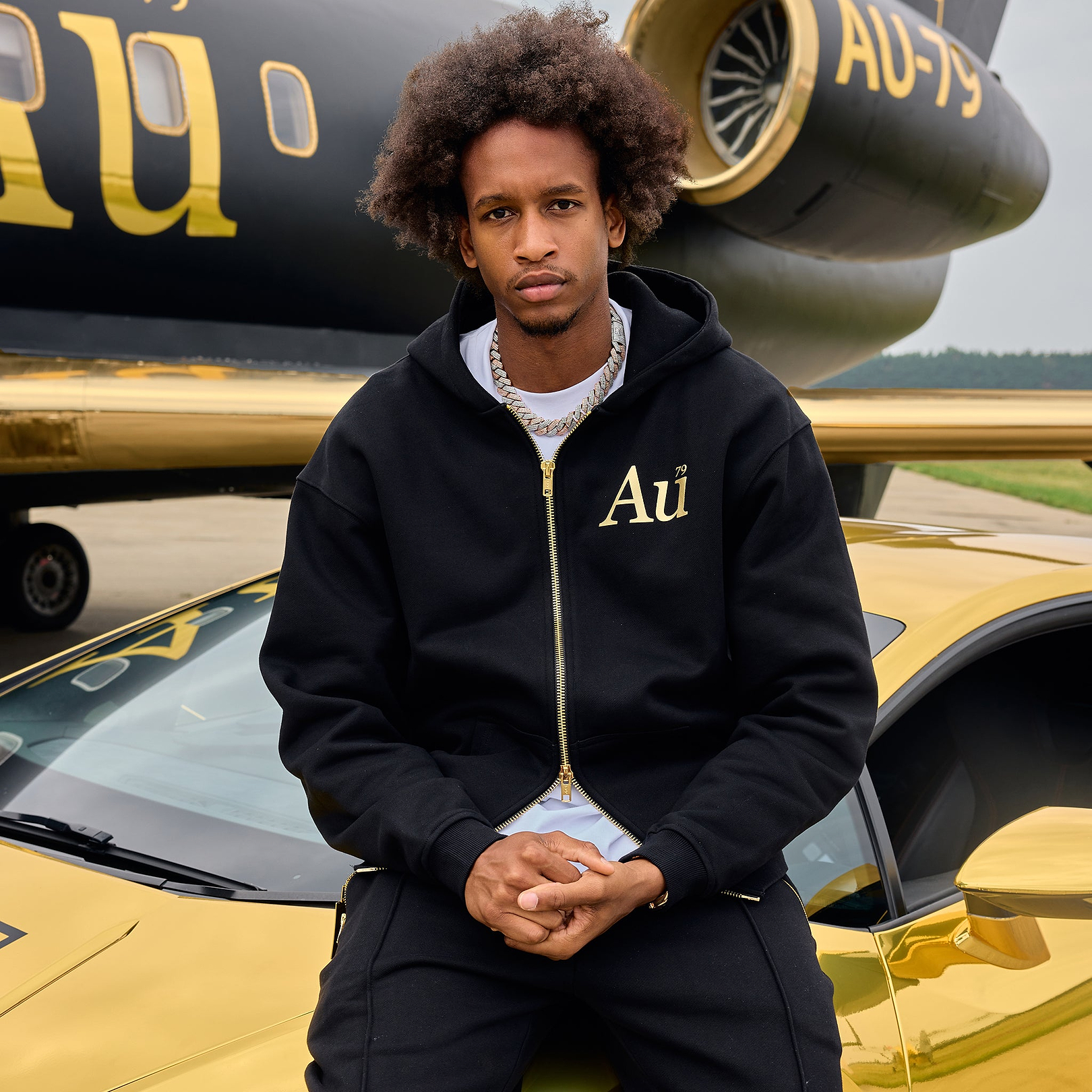 Man wearing black Au Vodka zip up hoodie sitting on a gold car in front of a private jet