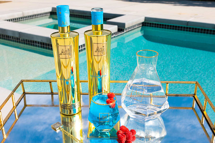 Two Au Vodka bottles, a blue drink with raspberries, and a clear carafe on a gold tray by a pool