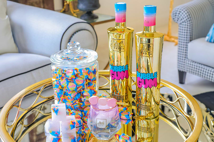 Two gold Au Vodka Bubblegum bottles, a jar of bubblegum, and a glass with pink candies on a gold tray