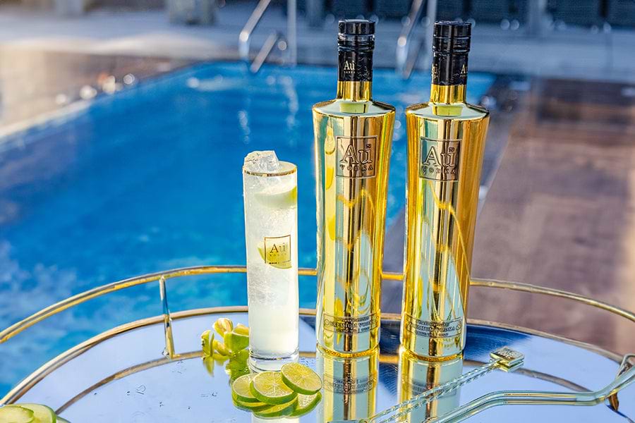 Two gold Au Vodka bottles and a cocktail with lime slices on a reflective tray by a pool