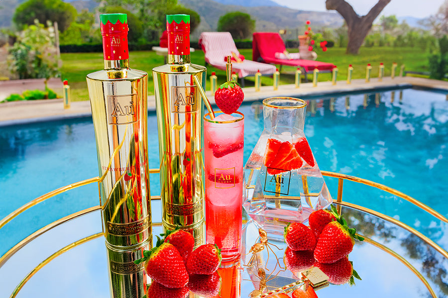Two Au Vodka Strawberry Blush bottles, a cocktail, and a carafe with strawberries by a pool