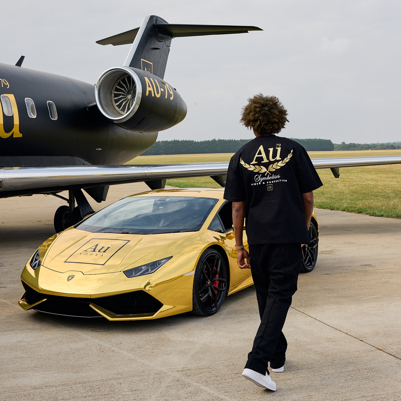 Person wearing black Au Vodka apparel walking toward a gold Lamborghini and private jet