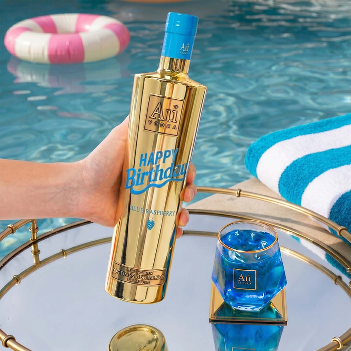 Hand holding Au Vodka USA 750ml Blue Raspberry Happy Birthday bottle next to a glass of blue drink by a pool