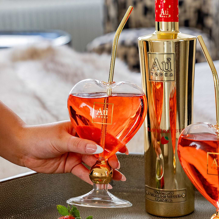 Hand holding heart shaped Au Vodka glass with gold straw next to gold bottle of Au Vodka Strawberry Burst