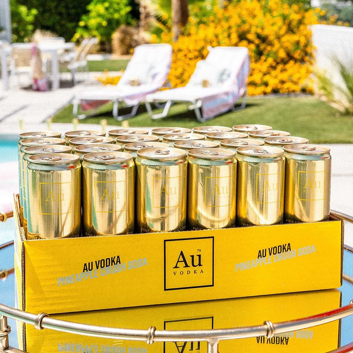 Case of Au Vodka Pineapple Crush Soda gold cans on a reflective surface outdoors