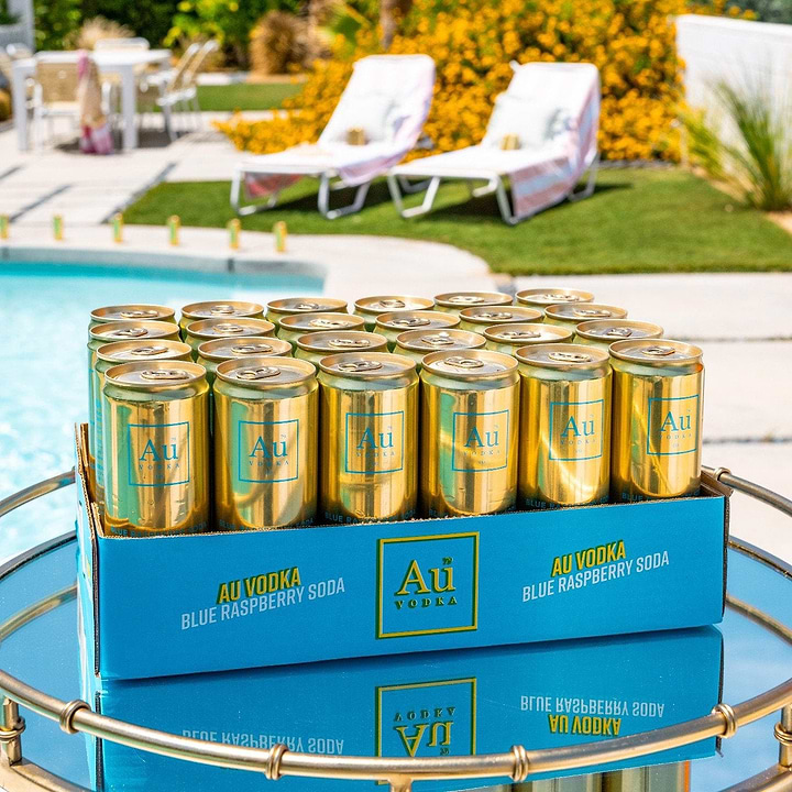 Case of Au Vodka Blue Raspberry Soda cans on a reflective table by a pool