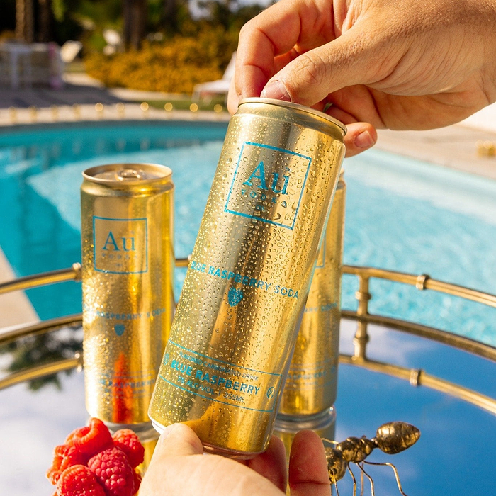 A hand holding an Au Vodka Blue Raspberry Soda can over a tray with other cans and raspberries