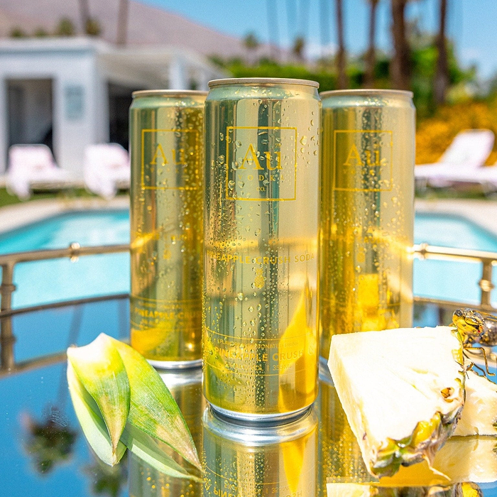 Three Au Vodka USA Pineapple Crush cans on a tray next to pineapple slices by a pool