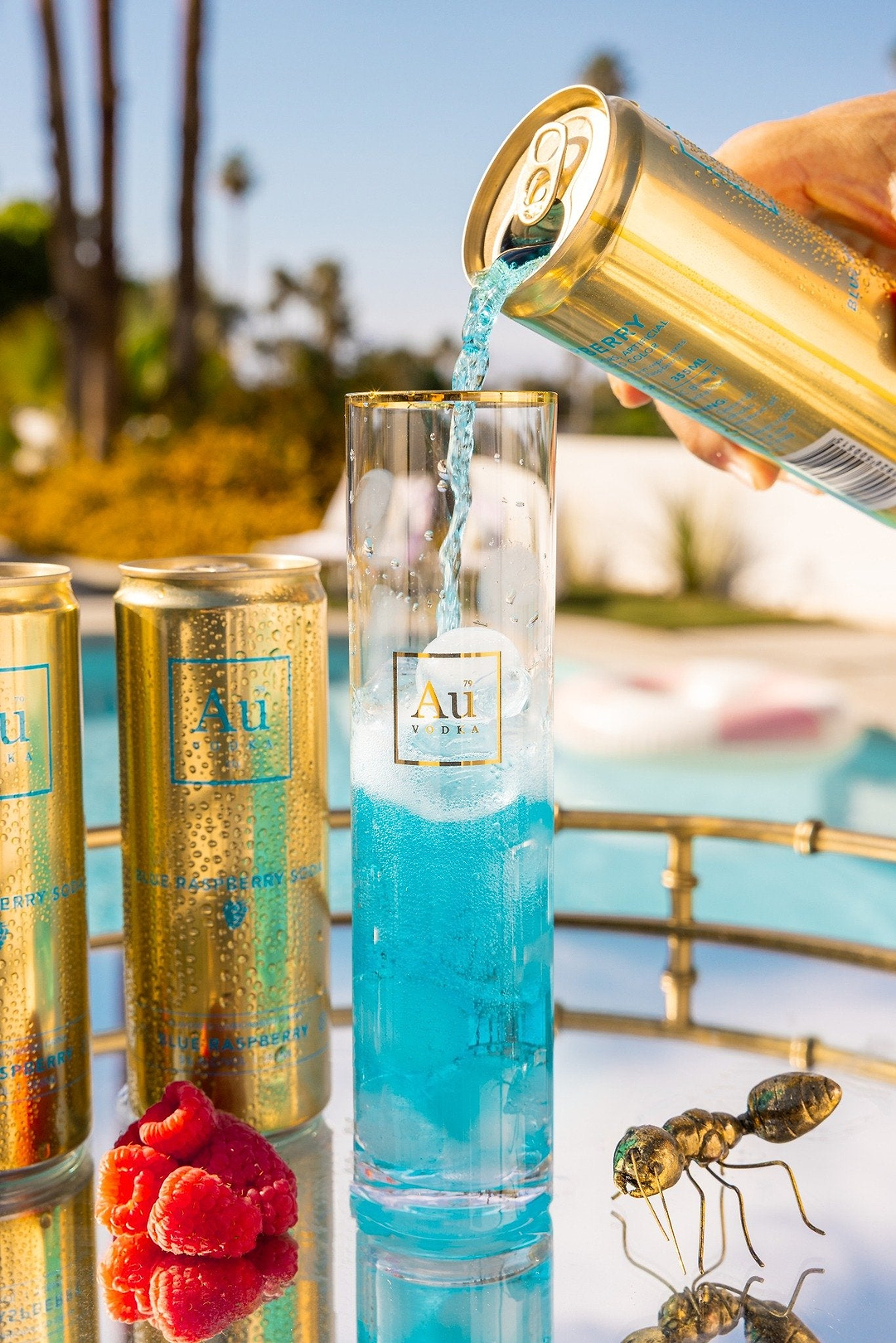 Au Vodka Blue Raspberry soda being poured into a tall glass with ice next to fresh raspberries