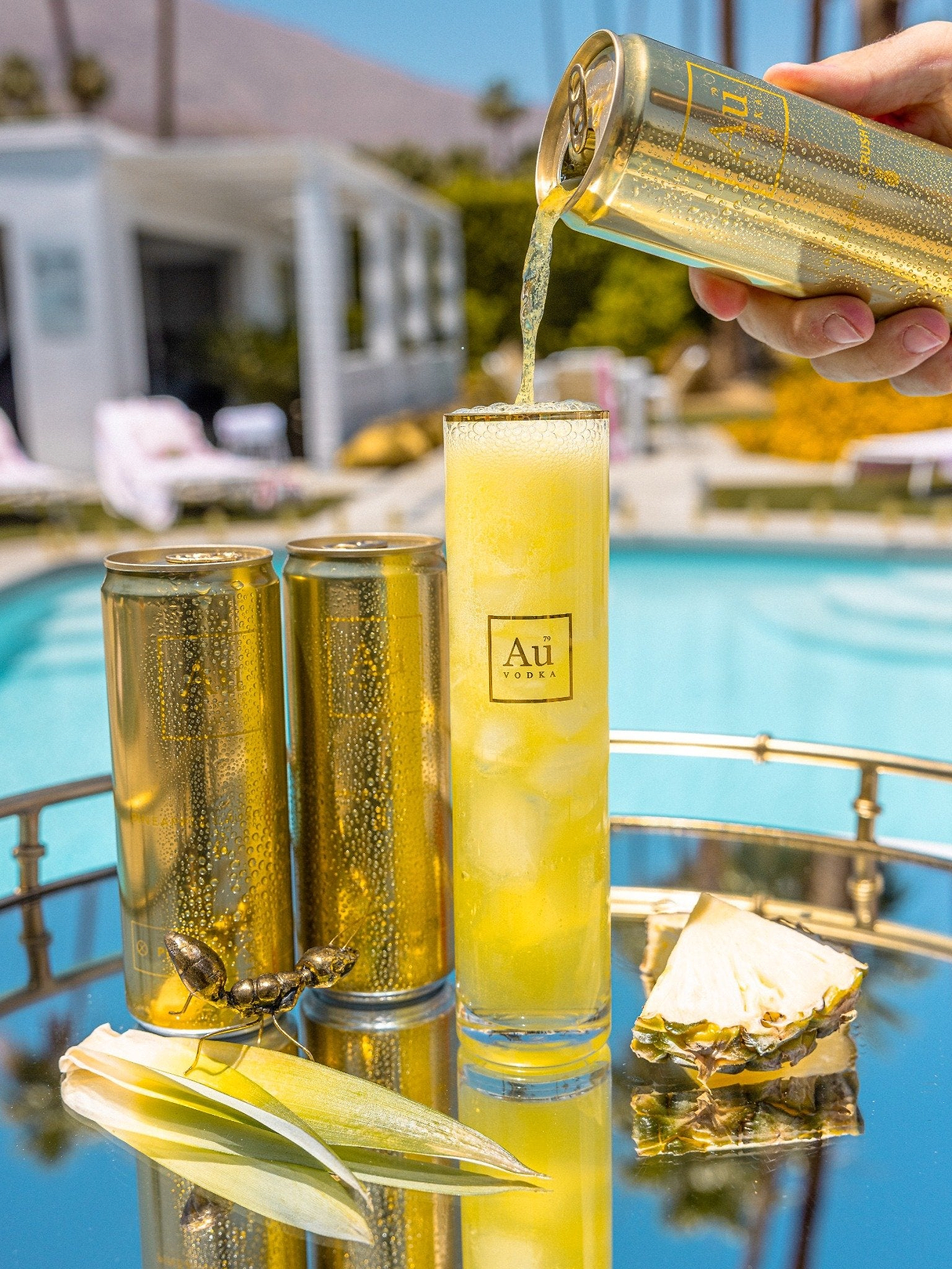 Au Vodka Pineapple Crush being poured from a gold can into a tall glass with ice by a swimming pool