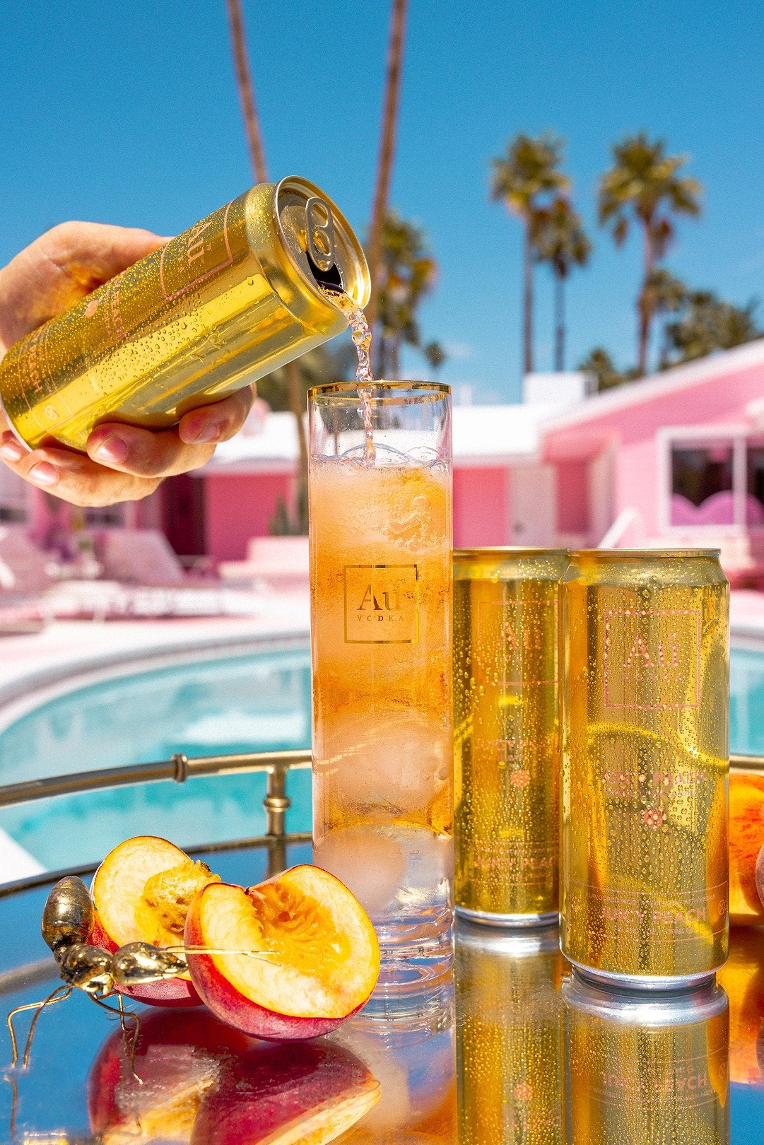 Au Vodka Juicy Peach being poured into a tall glass with ice beside fresh peach slices by a pool