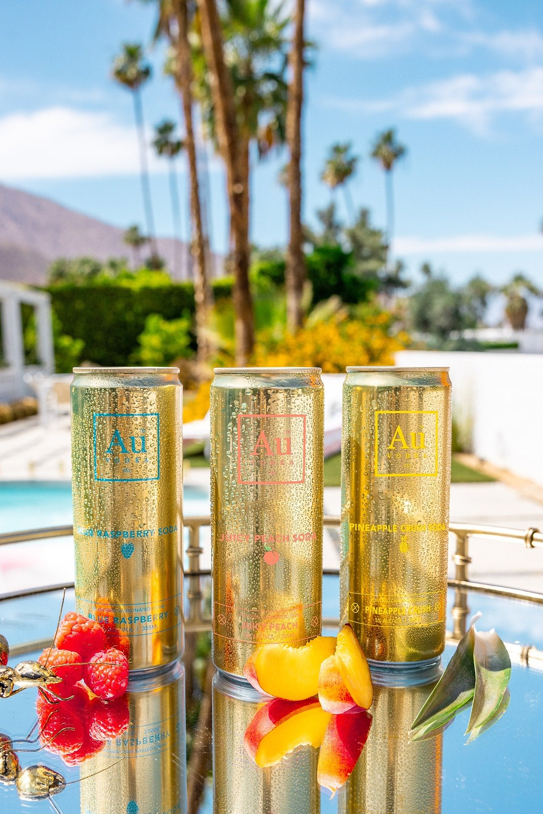 Three gold Au Vodka cans in Blue Raspberry, Juicy Peach, and Pineapple Crush flavors with fresh fruit on a tray