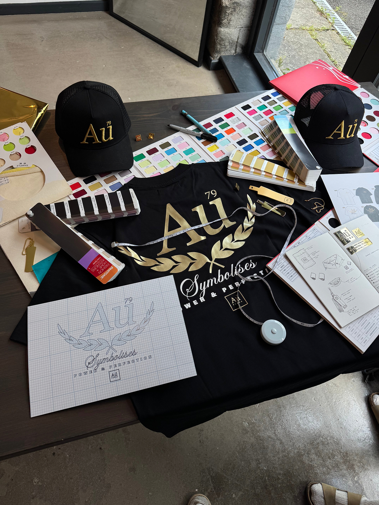 Au Vodka branded merchandise including black caps t-shirt design sketches and Pantone color swatches on a table