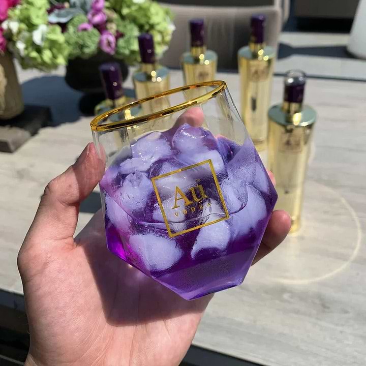 Hand holding a geometric Au Vodka glass filled with purple liquid and ice cubes with gold Au Vodka bottles in background
