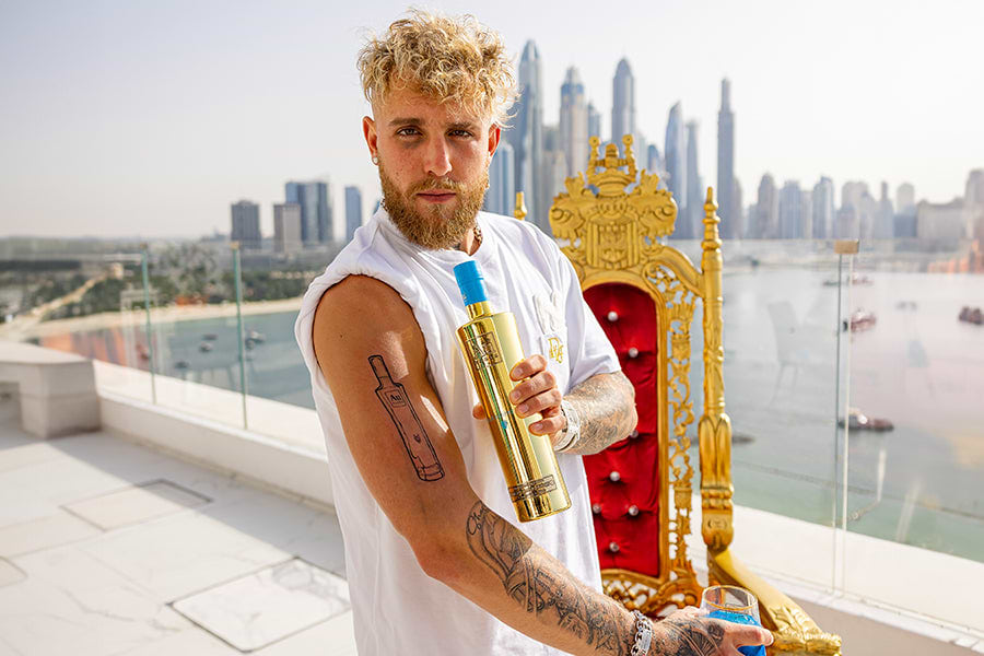 Jake Paul holding a gold Au Vodka bottle with a blue top, standing on a rooftop with a city skyline