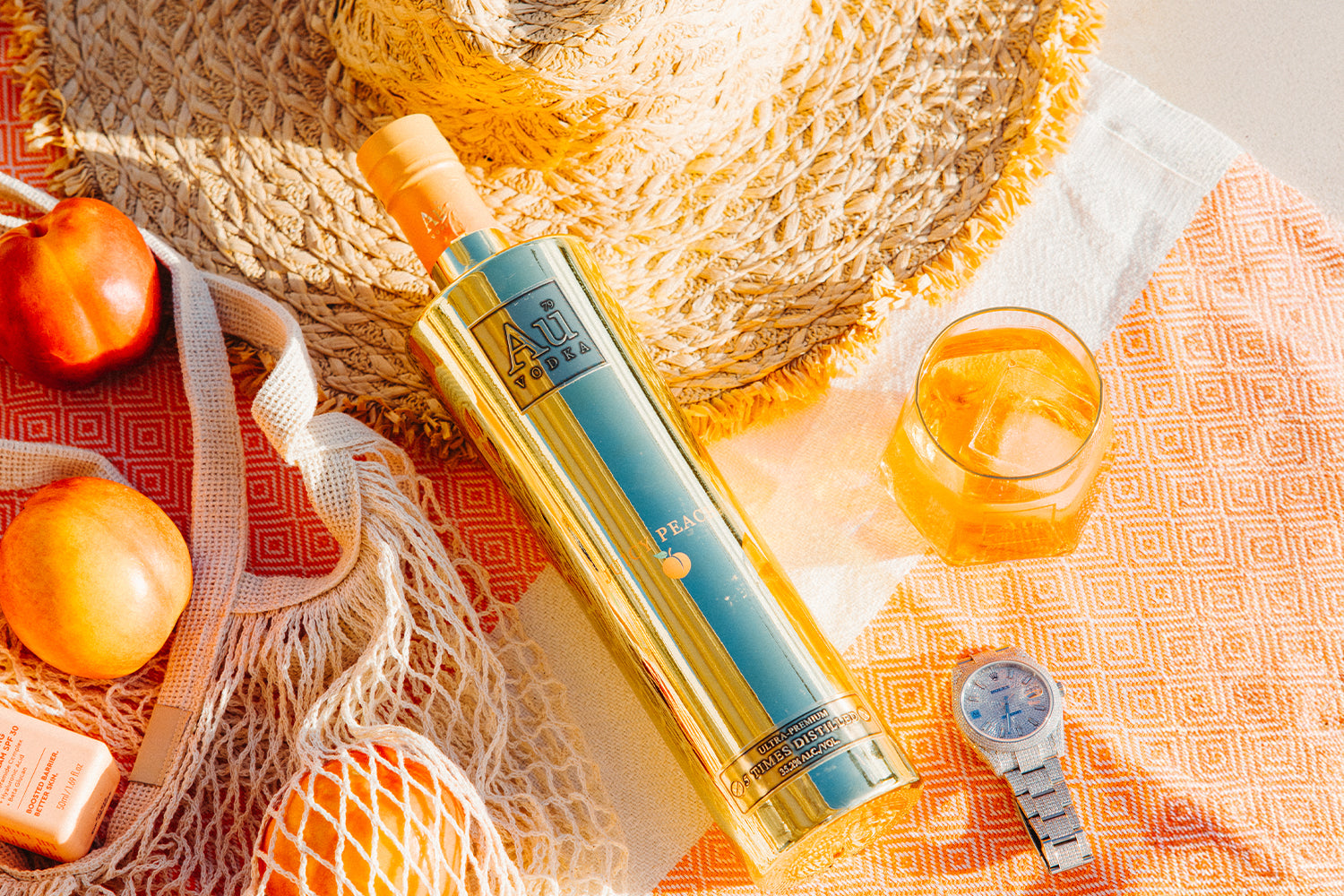 Au Vodka Peach bottle with a glass of drink, peaches, straw hat, and watch on a patterned beach towel