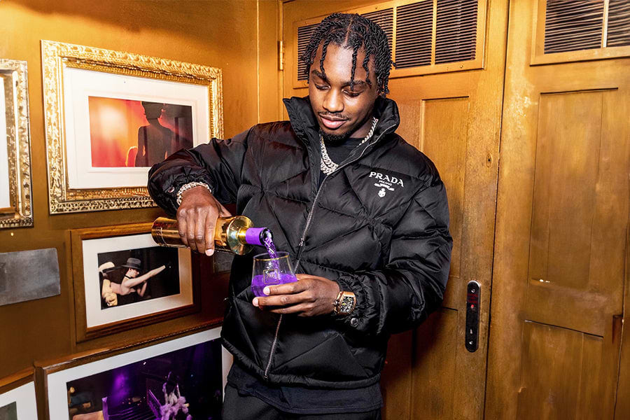 Man in black Prada puffer jacket pouring purple liquid from a gold bottle into a glass