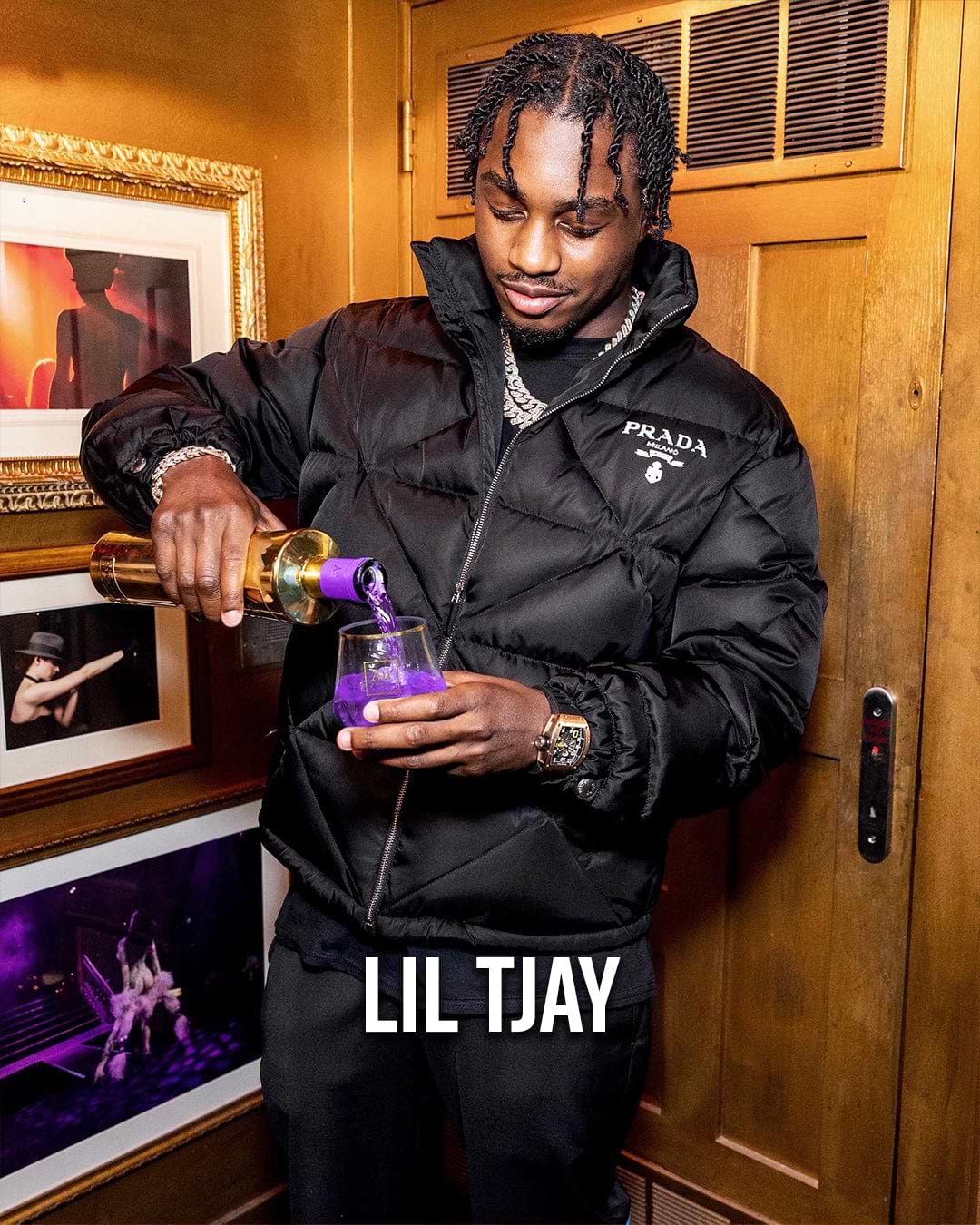 Lil Tjay pouring purple liquid into a glass, wearing a black Prada puffer jacket and diamond chains