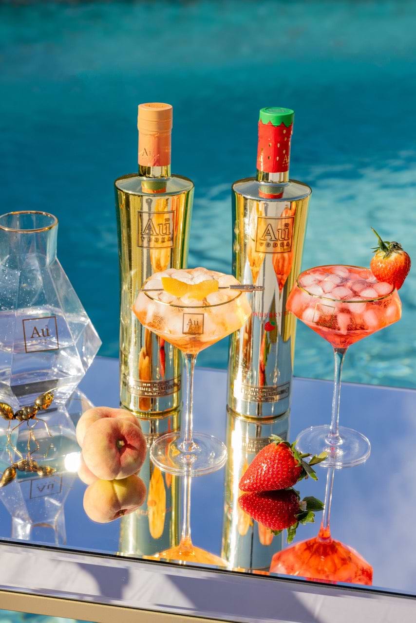 Au Vodka bottles with peach and strawberry cocktails on a mirrored surface by a pool