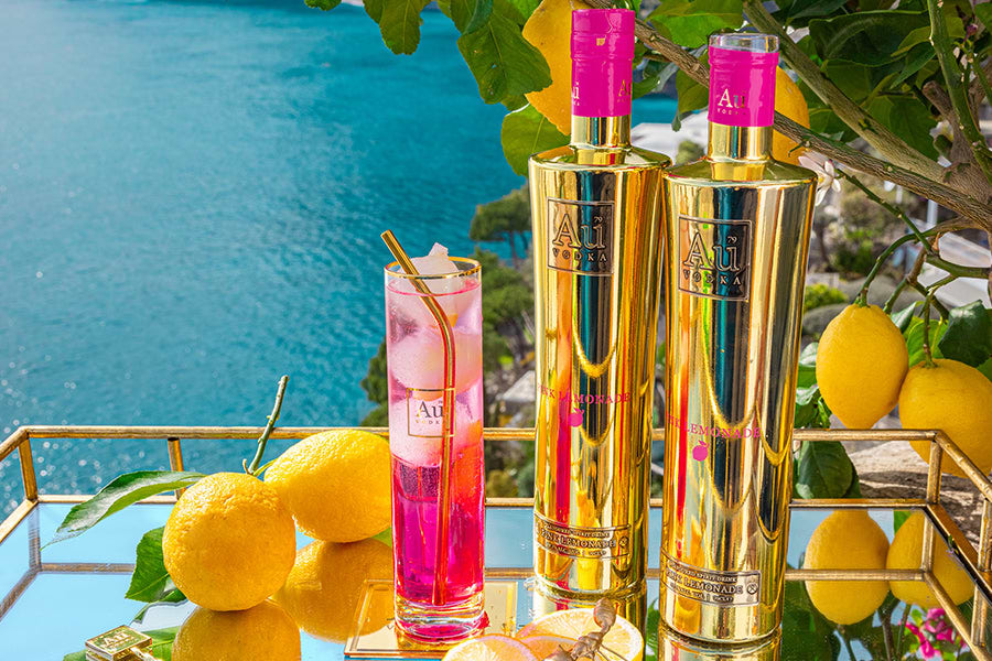 Two Au Vodka Pink Lemonade bottles and a cocktail on a mirrored tray with lemons and ocean background