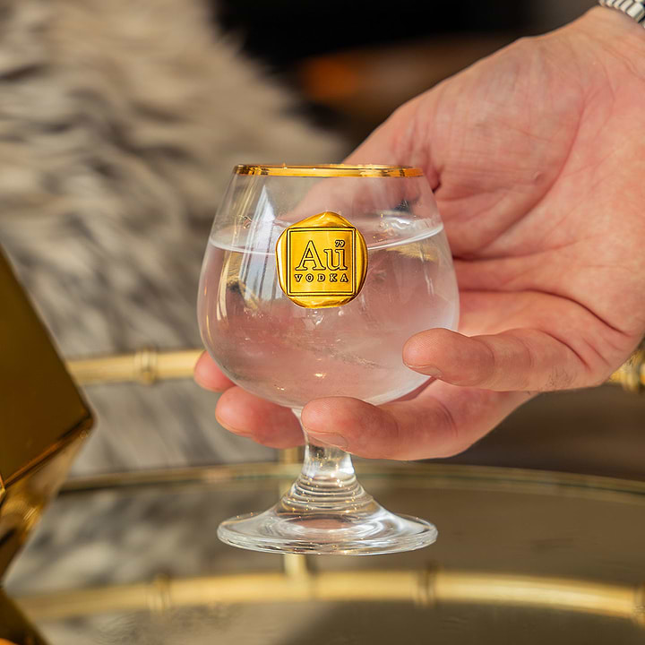 Hand holding an Au Vodka branded glass with a gold rim and gold emblem