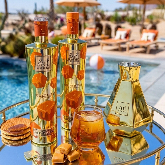 Two bottles of Sticky Toffee Vodka by Au Vodka on a reflective surface with a pool and lounge chairs in the background.