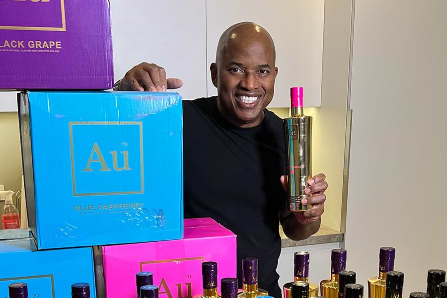 Man holding a gold bottle of Au Vodka with a pink top, surrounded by boxes of Au Vodka