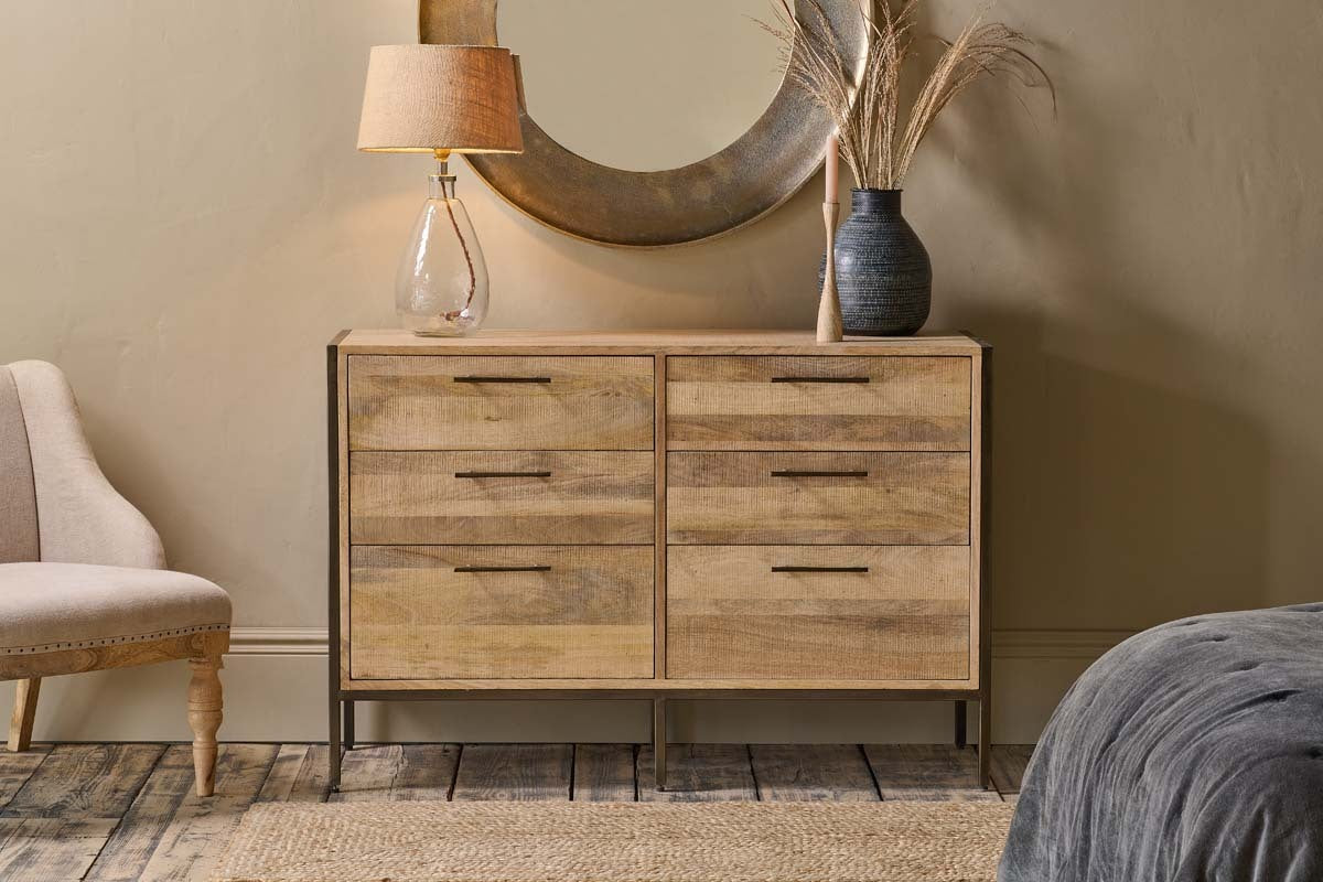 The Dasai Mango Wood Chest of Drawers by nkuku, featuring six compartments, stands against a beige wall. On top sit a glass lamp, round mirror, black vase with dried grasses; a woven rug lies below, with part of a cream chair and gray blanket visible.
