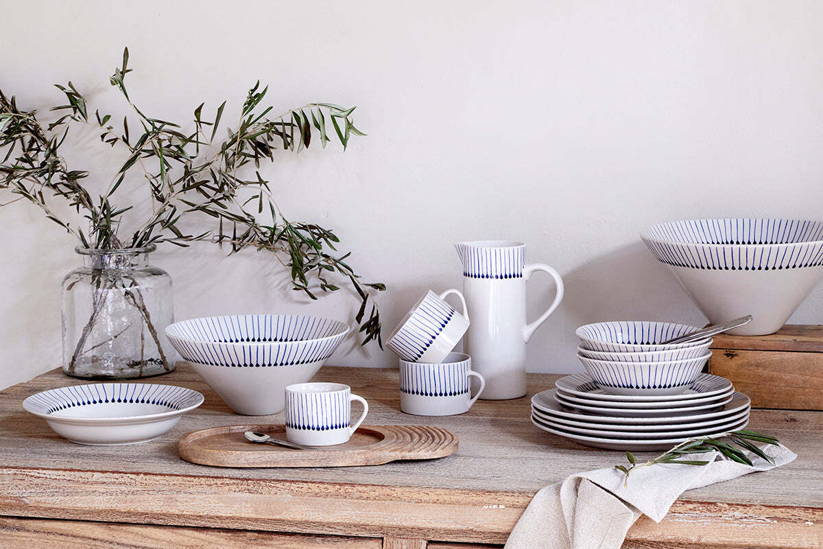 A rustic wooden table displays nkuku’s Iba Mug - Indigo - Large (Set of 2), hand-painted with thin blue stripes, alongside a pitcher, bowls, plates, leafy branches in a jar, a beige napkin, and a wooden board for a cozy minimalist look.