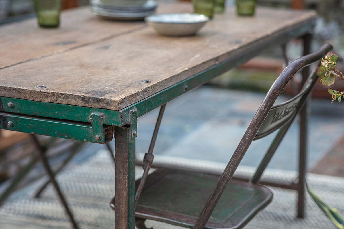 A close-up of the nkuku Ishan Reclaimed Folding Dining & Coffee Table with a weathered wood top and green metal frame, set on a patio with plates, green glasses, and an old metal folding chair amidst nearby plant leaves.