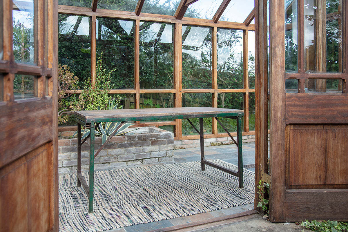 The nkuku Ishan Reclaimed Folding Dining & Coffee Table rests on a woven rug in a sunlit greenhouse, with open wooden doors framing vibrant greenery outside.