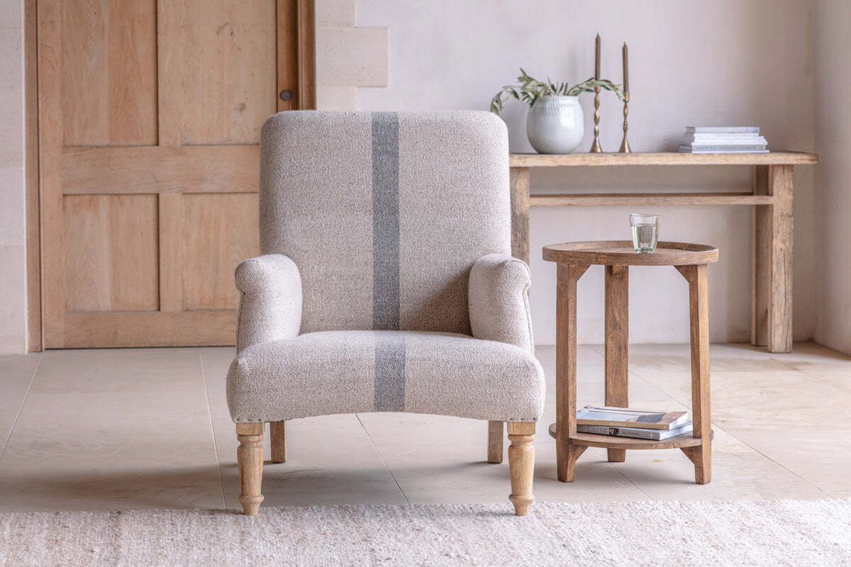 The Kallu Wool & Cotton Occasional Chair by nkuku, in natural beige with textured upholstery and gray stripes, sits beside a rustic wooden side table with a glass of water. A long wooden console table with books and decor is in the background.